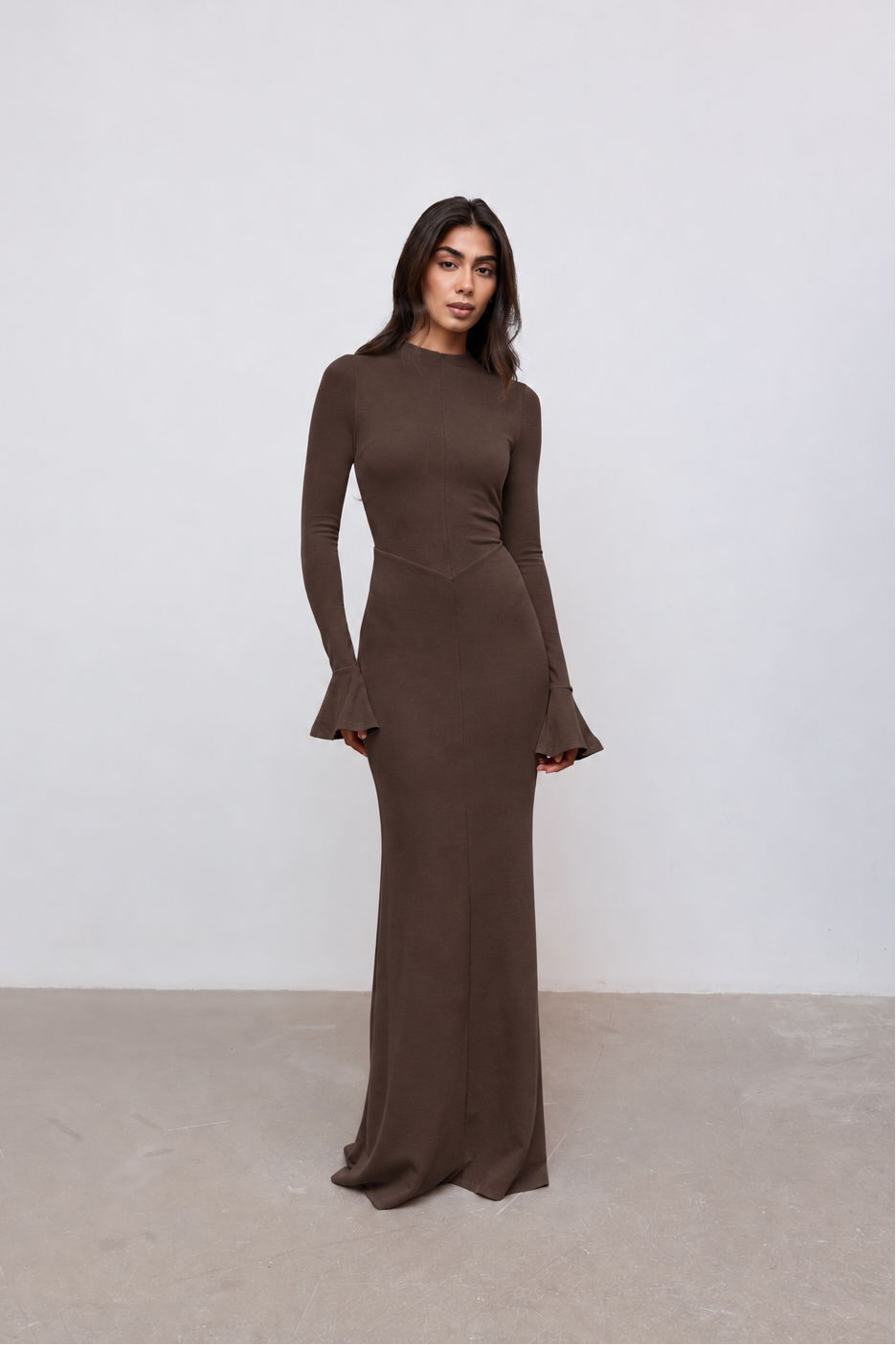 A woman modeling a long-sleeve, floor-length brown dress with flared cuffs, standing against a plain light gray wall and concrete floor.