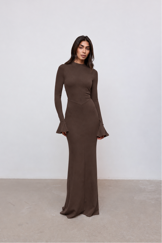 A woman modeling a long-sleeve, floor-length brown dress with flared cuffs, standing against a plain light gray wall and concrete floor.