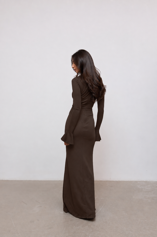 A woman wearing a long-sleeved, dark brown maxi dress, shown from the back in a minimalist studio setting with a white wall and light concrete floor.