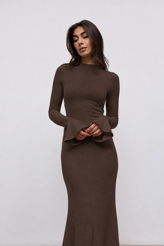 A woman modeling a long-sleeve, bodycon maxi dress in a dark brown color, featuring flared cuffs and a high neckline, set against a plain light gray background.